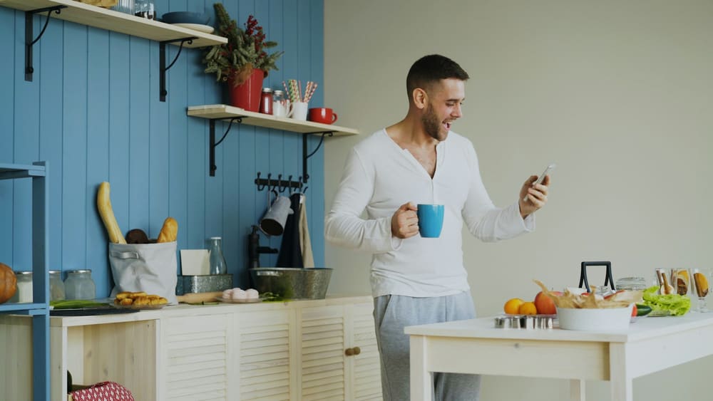 Un homme en cuisine, un café à la main, lit son téléphone en souriant