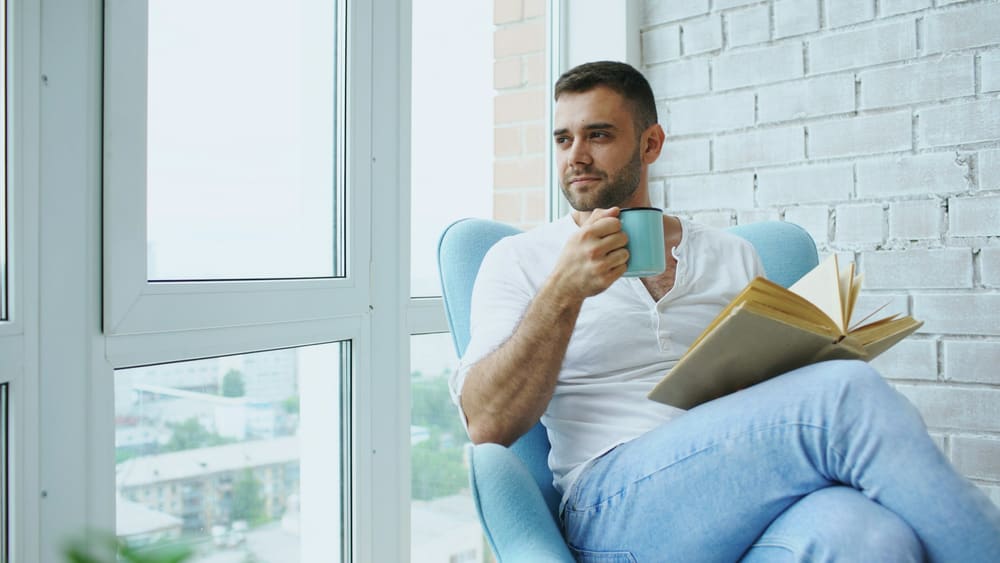Un homme lit dans un fauteuil, un café à la main, près de la fenêtre